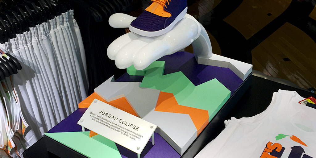 Image of a shoe displayed on a 3d cartoon-esque hand and a colorful cut-out base, with colors that match the colors of the shoe